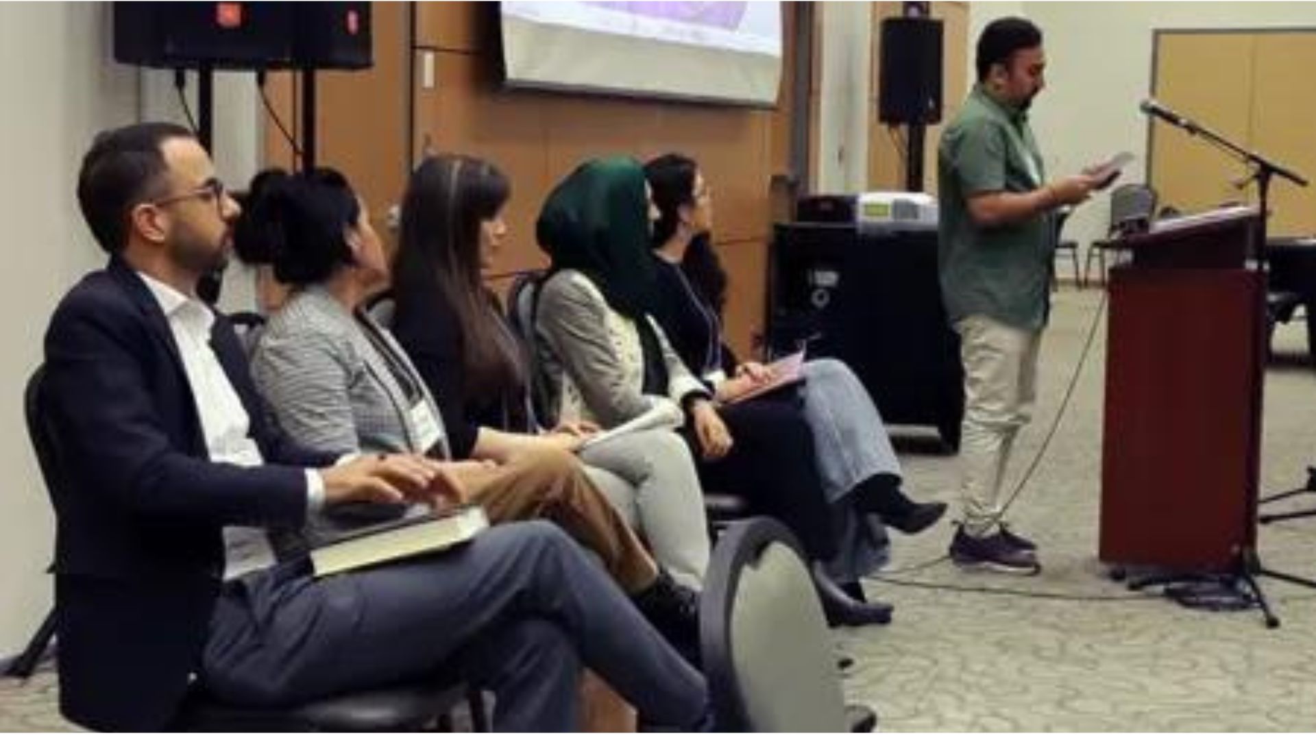 Five people sitting in a conference, listening to a speaker at a lectern.